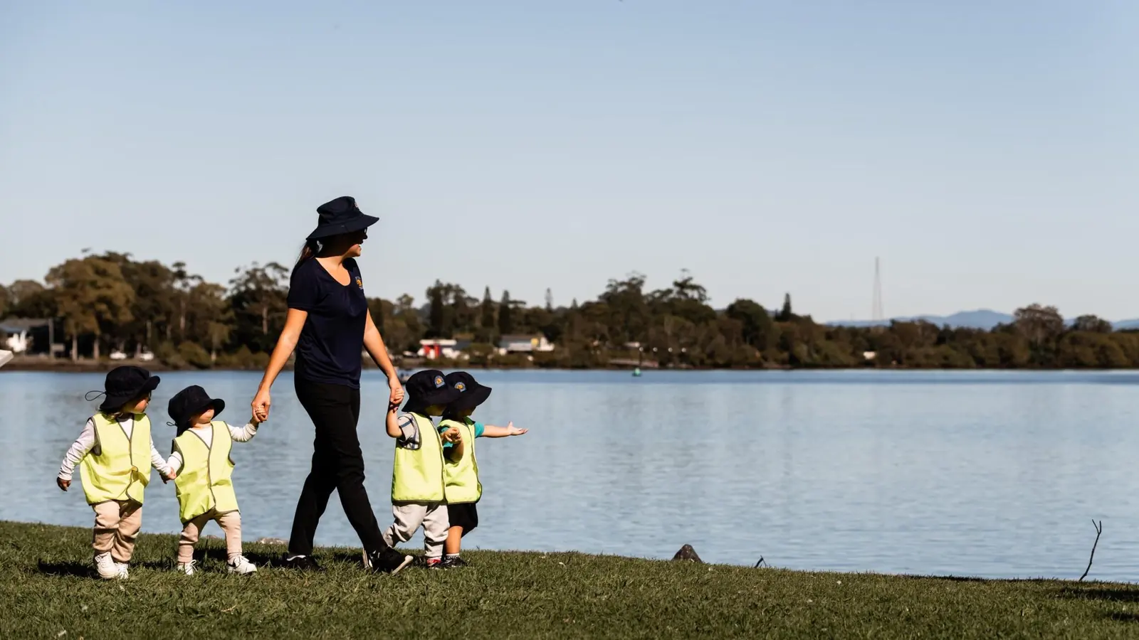 An educator walking with children along the grass beside the water